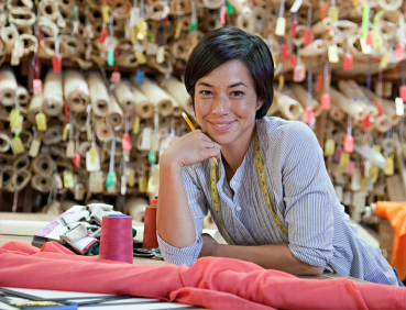 Smiling woman leaning on a desk with hundreds of rolls of fabric in the background.